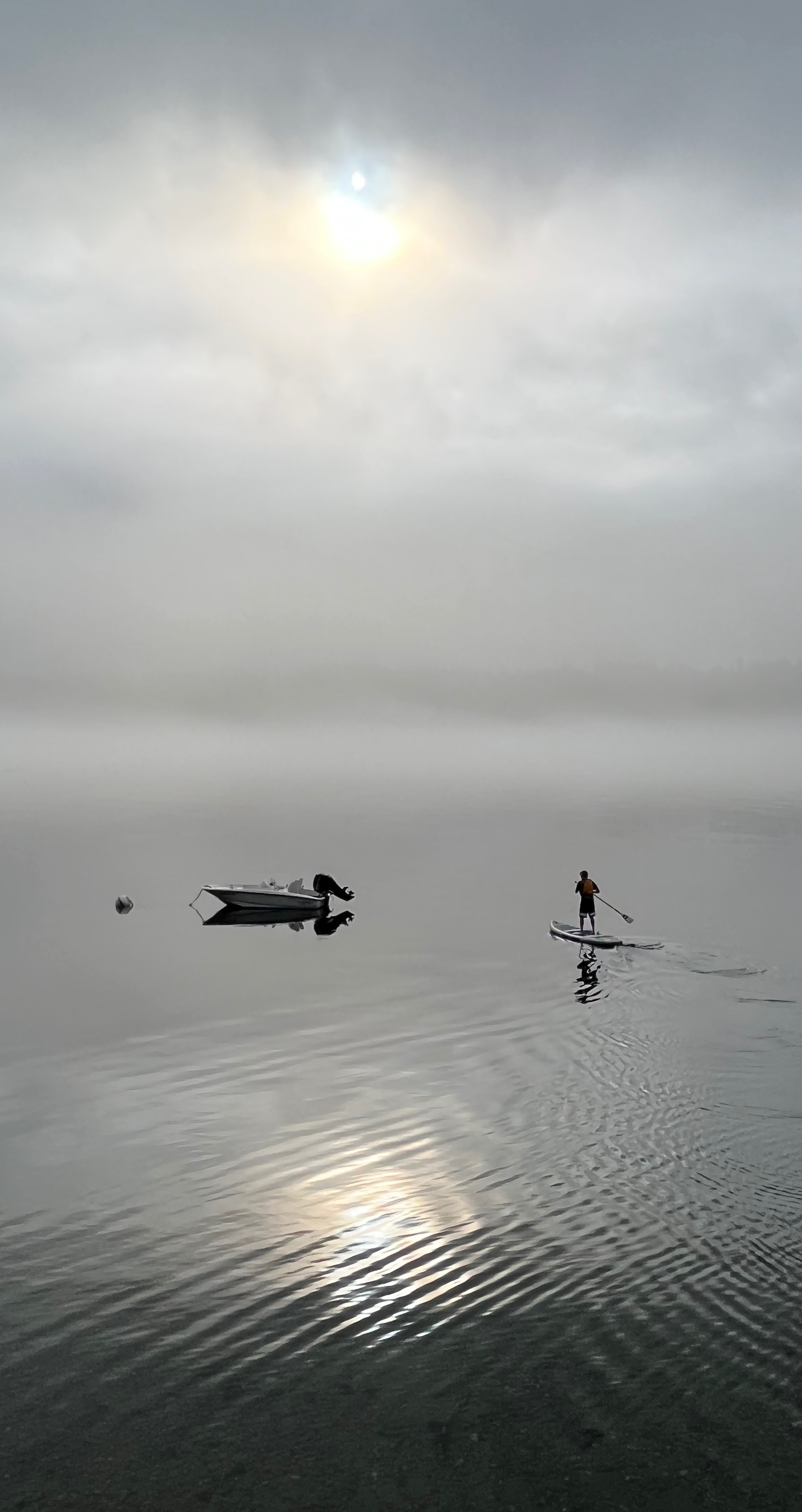 A paddleboarder on glassy, misty water with the sun breaking through clouds